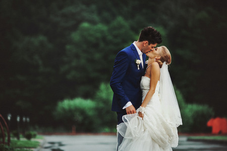 Groom with stylish hair kisses a tiny blonde bride in the parkの写真素材