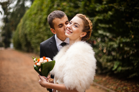 Groom kisses a delicate bride in a cheek while posing in the parkの写真素材