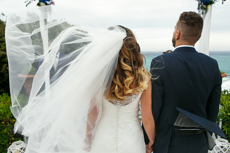 Wind blows bride's veil and groom's jacket during the ceremony by the seaの写真素材