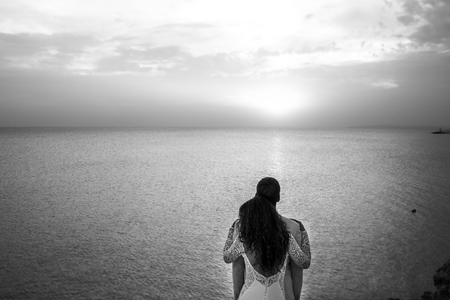 Brunette woman hugs her man from behind while standing on the rock over the seaの写真素材