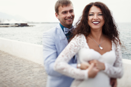 Groom holds bride's waist tightly while they pose by the seaの写真素材