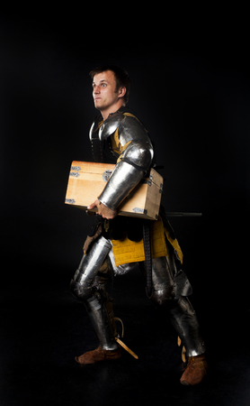 Humorous studio shot of young man dressed as medieval knight carrying away treasure chest (on black background)の写真素材