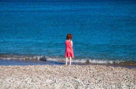 Little redhead girl in pink dress at seashore looking at the wavesの写真素材