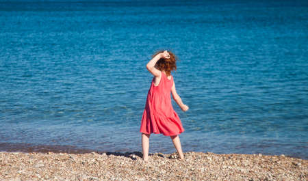 Little redhead girl in pink throwing pebbles at sea in mock pose of ancient athleteの写真素材