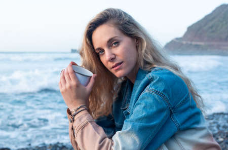 Portrait of a young blonde woman on the beach. She is wearing a blue denim jacket, holding a mug looking at the cameraの写真素材