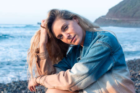 Portrait of a young blonde woman on the beach. She is wearing a blue denim jacket, holding her hair looking at cameraの写真素材