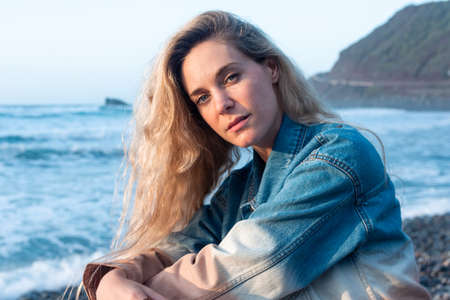 Portrait of a young blonde woman on the beach. She is wearing a blue denim jacket, looking at cameraの写真素材