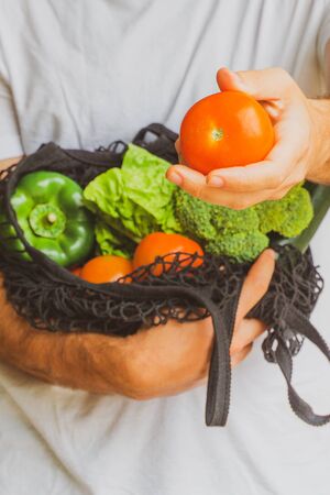 Hand of a Man showing a tomato and holding a cotton mesh bag reusable full of eco vegetables on the background. Eco friendly lifestyle and no plastic concept.の写真素材