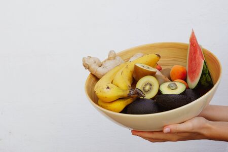 Two woman hands holding a bowl full of summer fruits isolated on white. Bowl full of banana, kiwi, avocado, nuts and watermelon. Healthy food concept.の写真素材