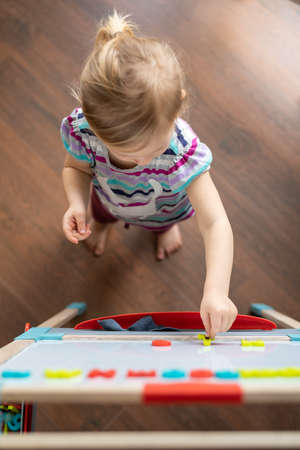 a 2-year-old child stands on a brown parquet and lays out magnetic numbers on the board with interest. View from aboveの写真素材