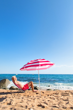 Senior man sun bathing at the beach under parasol with waves, sea and sandの写真素材