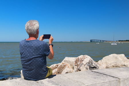Senior man sitting at the coast and taking a photo from bridge to mainland island Ile the Reの写真素材