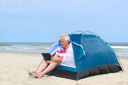 Single senior man with digital tablet camping in shelter at the beachの写真素材