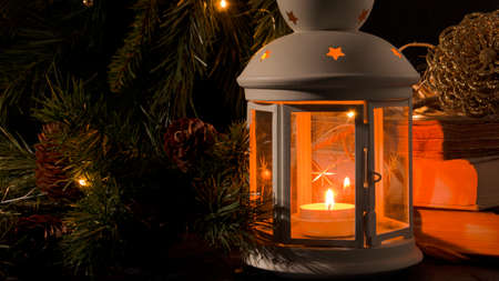 White lantern with a burning candle. Books, christmas tree branch and lights on background. Night dark picture. Christmas mystery mood.の写真素材