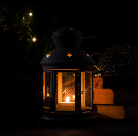 White candle lantern with burning candle. Blurred spruce brunch and garland lights on the dark background. Falling snow. Retro style picture.の写真素材