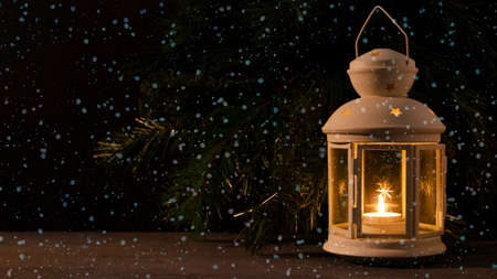 White candle lantern with burning candle. Blurred spruce brunch on the dark background. Falling snow. Retro style picture.の写真素材