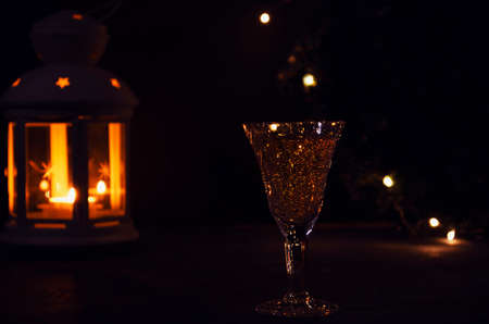 Glass with white wine with garland lights and candle lantern on the background. dark picture. Burning candle. Golden beverage in the glass. Beautiful lights reflected in the glass.の写真素材