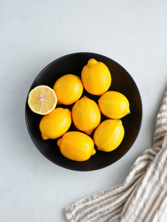 A black bowl holds several vibrant yellow lemons with a halved lemon placed nearby. The setting has a clean, light backdrop that enhances the freshness of the lemonsの素材