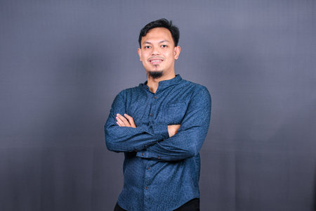 Smiling Asian man with blue shirt standing with hands folded isolated on grey background. Concept of engineering jobsの写真素材