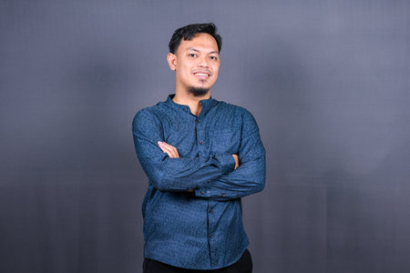 Smiling Asian man with blue shirt standing with hands folded isolated on grey background. Concept of engineering jobsの写真素材