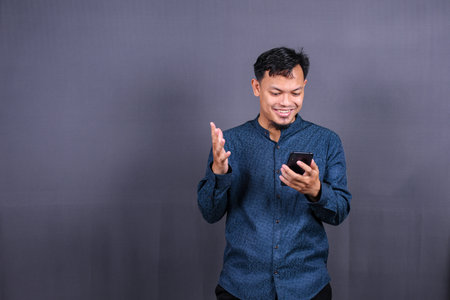 Happy young man in blue shirt posing isolated on gray background studio portrait. People lifestyle concept. Mock up copy space. Hold mobile phone, doing winner gestureの写真素材