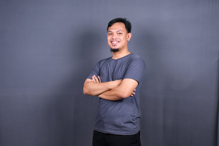 Smiling handsome asian man in gray t-shirt standing with crossed arms isolated on gray backgroundの写真素材
