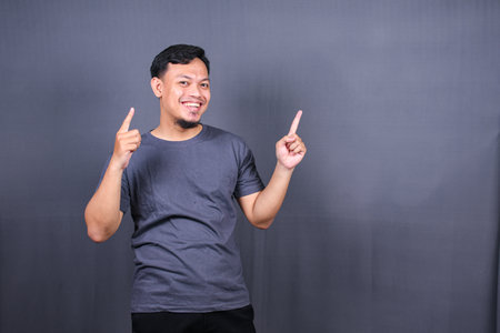 Young attractive smiling asian man in tshirt posing isolated on gray background, Mock up copy space. Pointing finger asideの写真素材