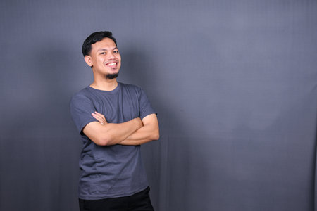 Smiling handsome asian man in gray t-shirt standing with crossed arms isolated on gray backgroundの写真素材