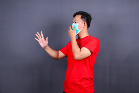 side view asian man in a medical mask show stop hand gesture to the coronavirus epidemic, covid-19. Concept of stop panic and fight against the virus. Wearing a red t-shirt on a grの写真素材