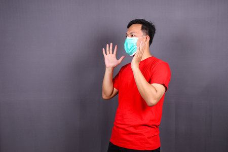 side view asian man in a medical mask show stop hand gesture to the coronavirus epidemic, covid-19. Concept of stop panic and fight against the virus. Wearing a red t-shirt on a grの写真素材
