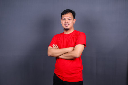 Portrait of attractive asian man in red t-shirt standing with crossed arms isolated on gray backgroundの写真素材