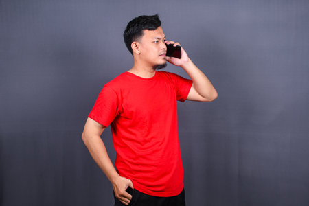Portrait of a young cheerful asian man wearing red t-shirt standing isolated over gray background, using mobile phoneの写真素材