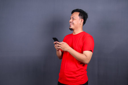 Portrait of a young cheerful asian man wearing red t-shirt standing isolated over gray background, using mobile phoneの写真素材
