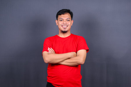 Portrait of attractive asian man in red t-shirt standing with crossed arms isolated on gray backgroundの写真素材