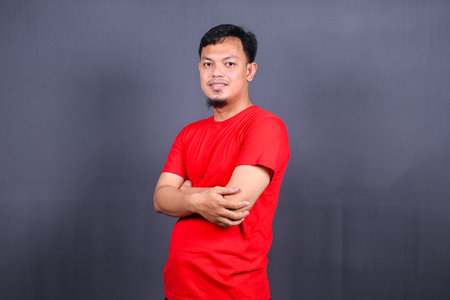 Portrait of attractive asian man in red t-shirt standing with crossed arms isolated on gray backgroundの写真素材