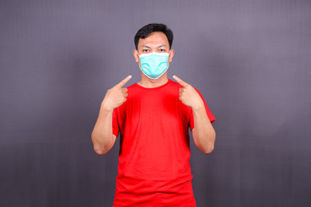 Asian young man pointing at face mask, asking to use measures against covid-19,  isolated on gray backgroundの写真素材