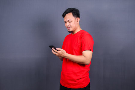 Portrait of a young cheerful asian man wearing red t-shirt standing isolated over gray background, using mobile phoneの写真素材