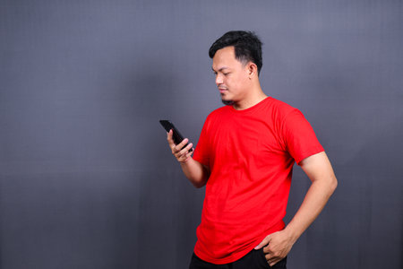 Portrait of a young cheerful asian man wearing red t-shirt standing isolated over gray background, using mobile phoneの写真素材