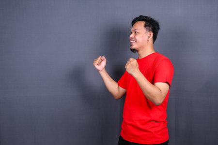 Portrait of an excited asian man standing with raised hands and looking at camera isolated on gray background celebrating succes, victory.の写真素材