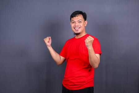 Portrait of an excited asian man standing with raised hands and looking at camera isolated on gray background celebrating succes, victory.の写真素材