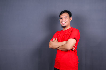 Portrait of attractive asian man in red t-shirt standing with crossed arms isolated on gray backgroundの写真素材
