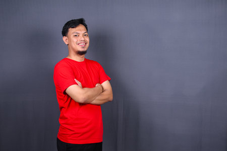 Portrait of attractive asian man in red t-shirt standing with crossed arms isolated on gray backgroundの写真素材