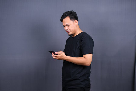 Photo of attractive asian guy holding mobile phone wear black t-shirt isolated gray color backgroundの写真素材