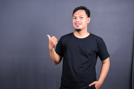 Portrait of a happy young asian man in black t-shirt pointing fingers away at copy space isolated over gray backgroundの写真素材