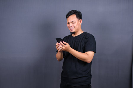 Photo of attractive asian guy holding mobile phone wear black t-shirt isolated gray color backgroundの写真素材