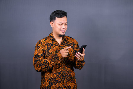 Portrait of asian young man wearing batik using cellphone on gray backgroundの写真素材