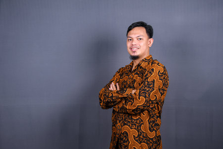 Portrait of attractive young asian man in batik indonesia standing with crossed arms isolated on gray backgroundの写真素材