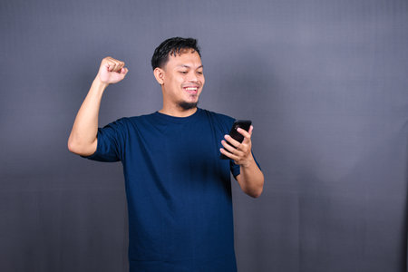 Young asian man using mobile phone and celebrating success on gray background
