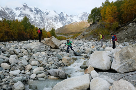 Svaneti, Georgia - circa October 2019: A group of tourists crossing stones along a riverbed Khaldechala. Snowy peaks Dzhangi-Tau and Shkhara with Khalde Glacier in foreground. Greater Causasusのeditorial素材
