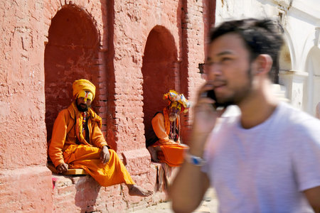 Kathmandu, Nepal - circa November 2019: Two Hindu holy men in traditional orange dress and a tourist passing by them at Pashupatinath Temple, Kathmandu, Nepal, Asiaのeditorial素材